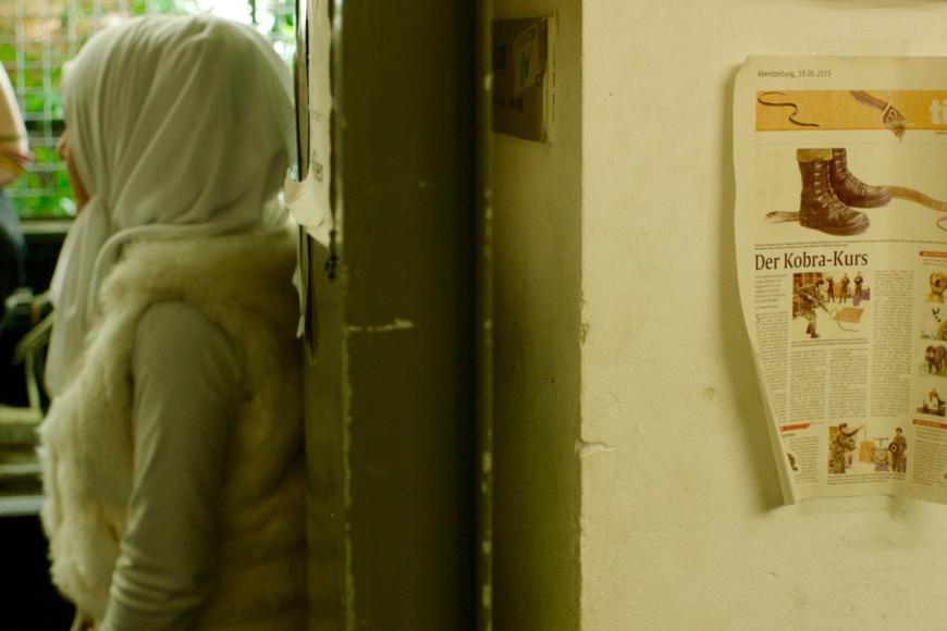 Eine Frau mit Kopftuch steht in einem Flur. Ein Zettel mit einem Zeitungsausschnitt hängt an der Wand.