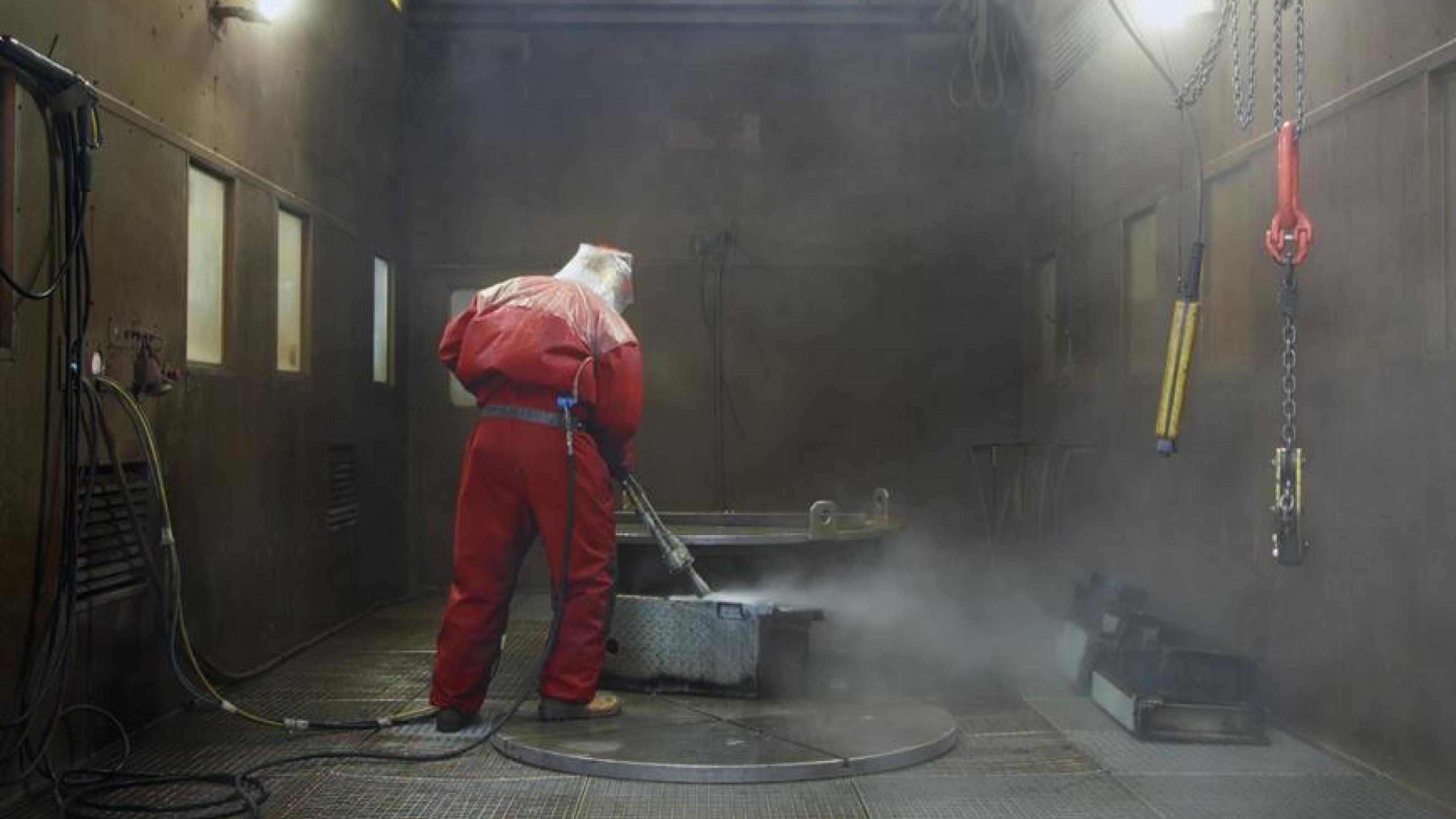 Ein Mann in einem roten Schutzanzug reinigt mit einen Hochdruckreiniger einen Metallkasten.