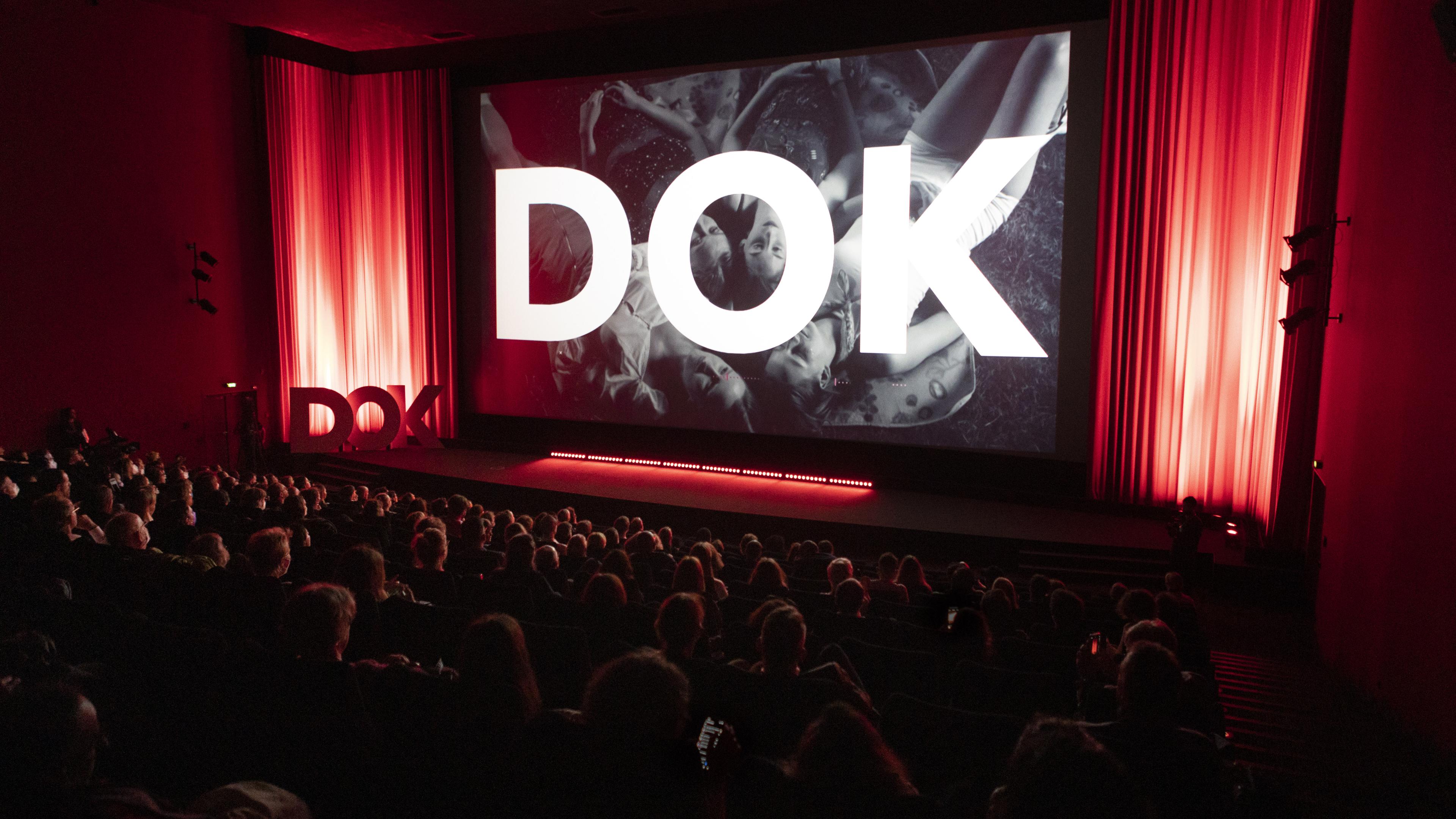 In einem Kinosaal mit vollbesetzten Reihen ist auf der Leinwand riesengroß das Wort Dok zu sehen
