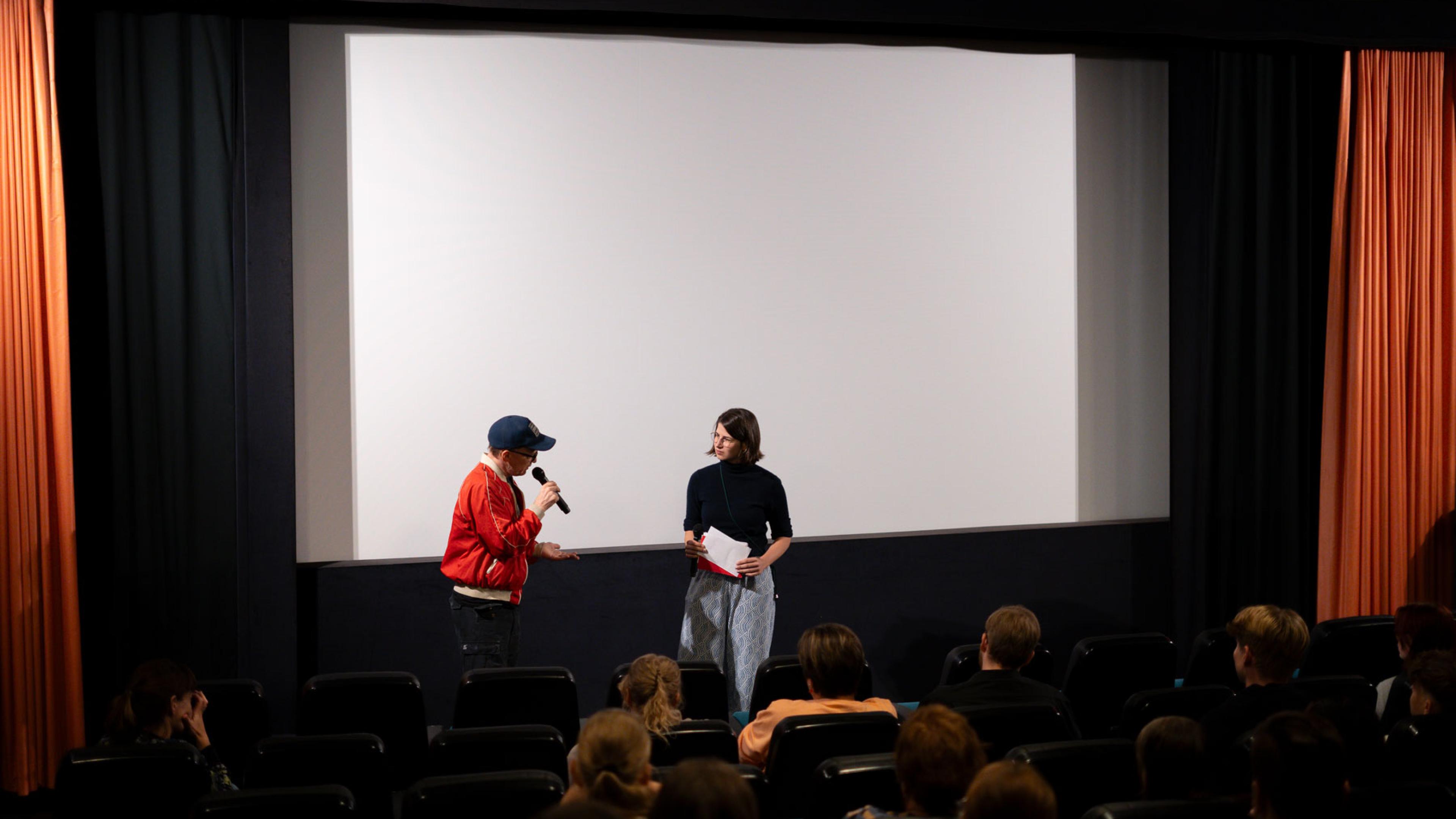 Tina Jany, DOK Bildung Koordinatorin im Filmgespräch mit einer Regisseurin in einem Kinosaal voller Kinder im Publikum