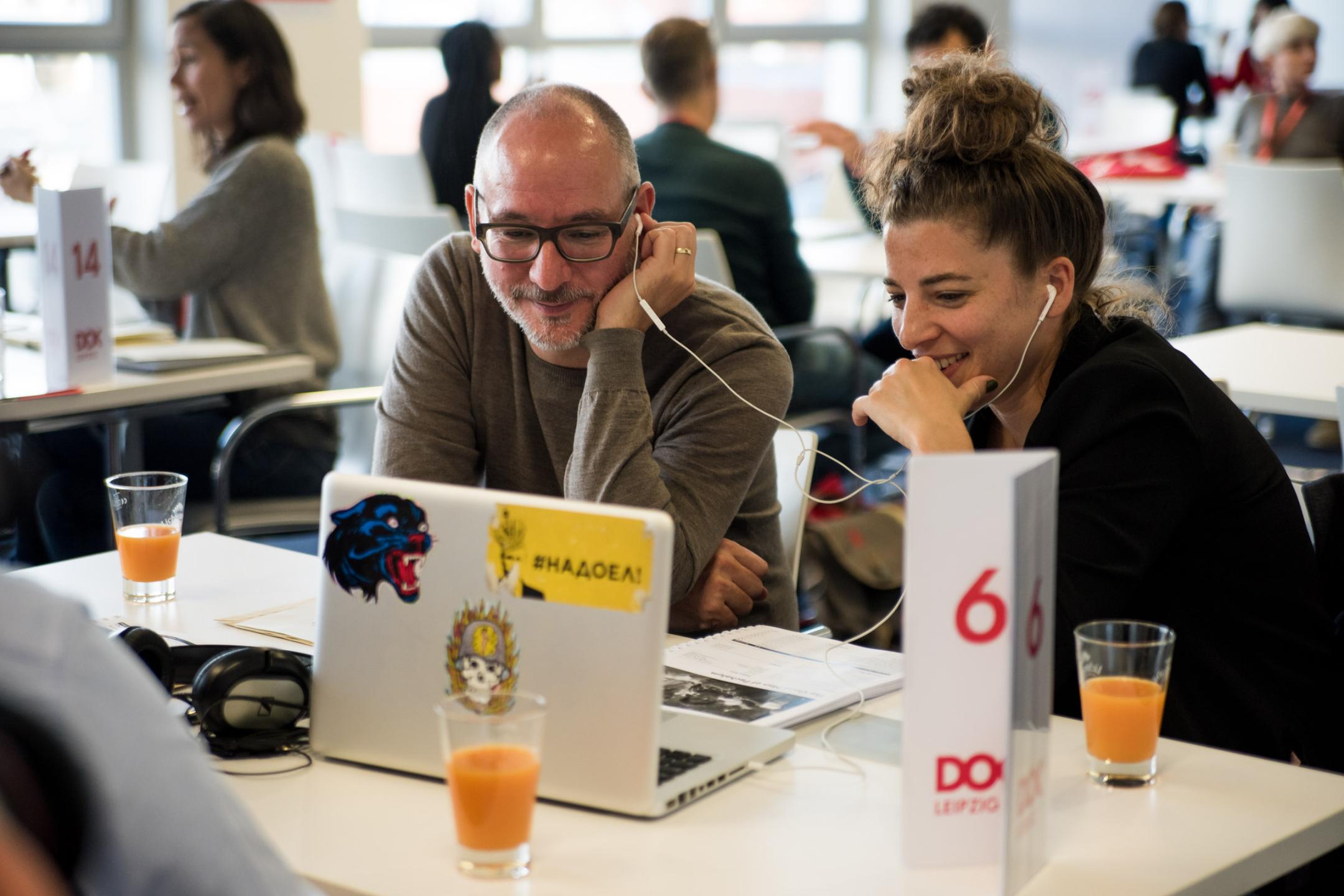 A man (left) and a woman (right) both with earphones sitting in front of a laptop that they share, watching at the display and smiling.