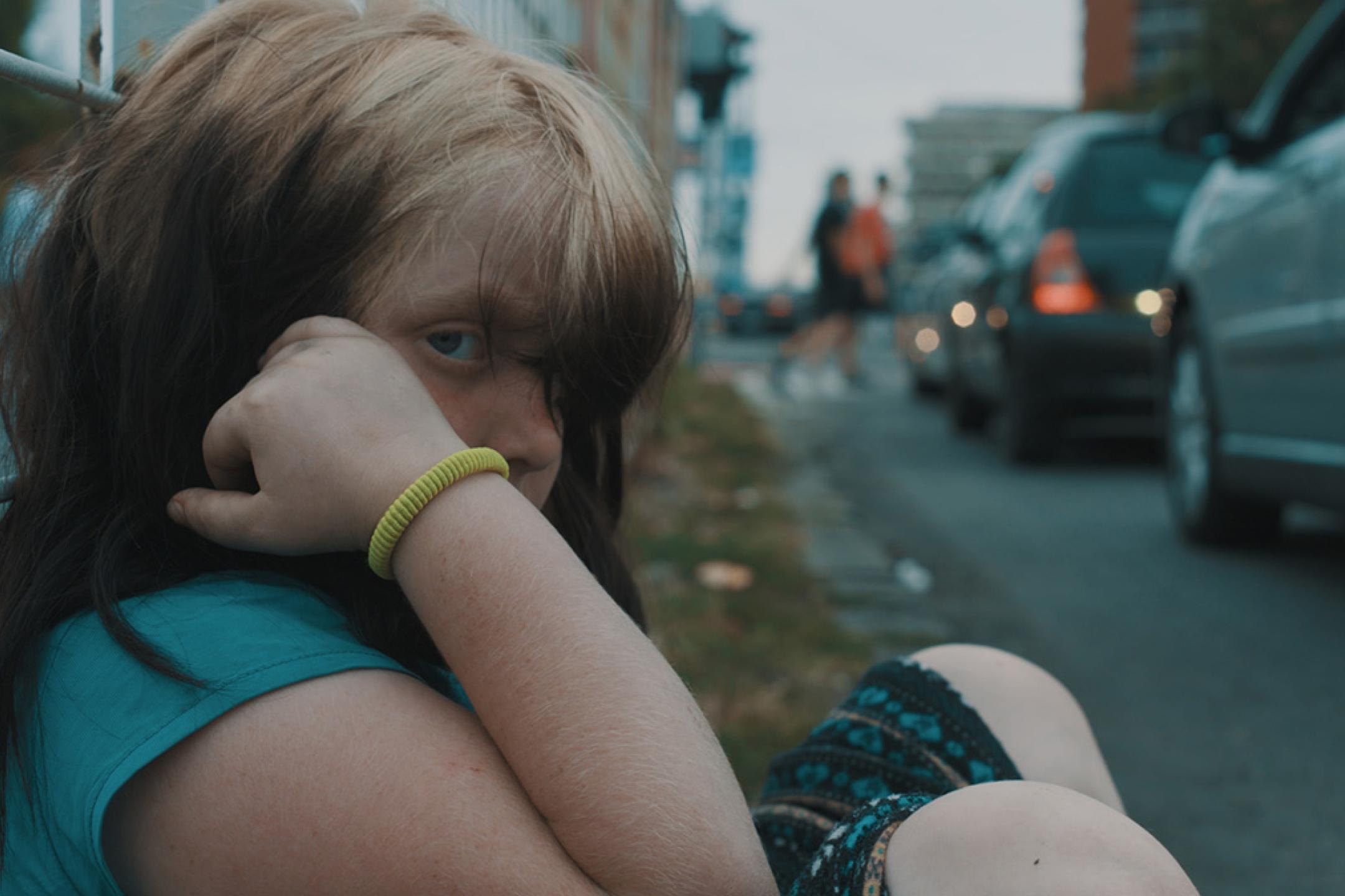 A young girl in summer clothes is sitting on the pathway at the side of a street. Her long hair almost covers her face. She pulls some hair aside to look with one eye into the camera.