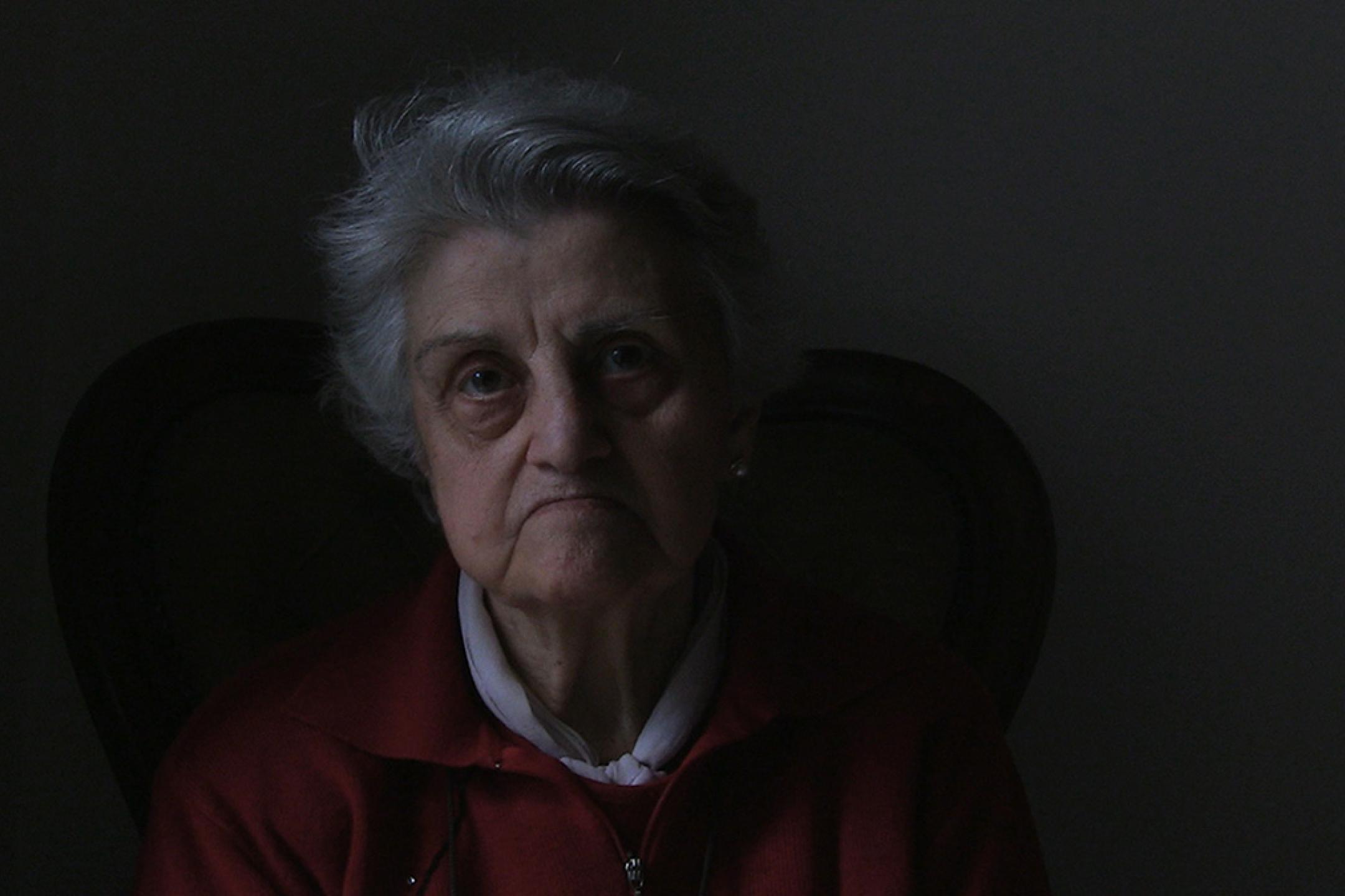 Porträt of an old woman. She looks straight and seriously into the camera, wearing shot white hair, a white shirt and a red cardigan.