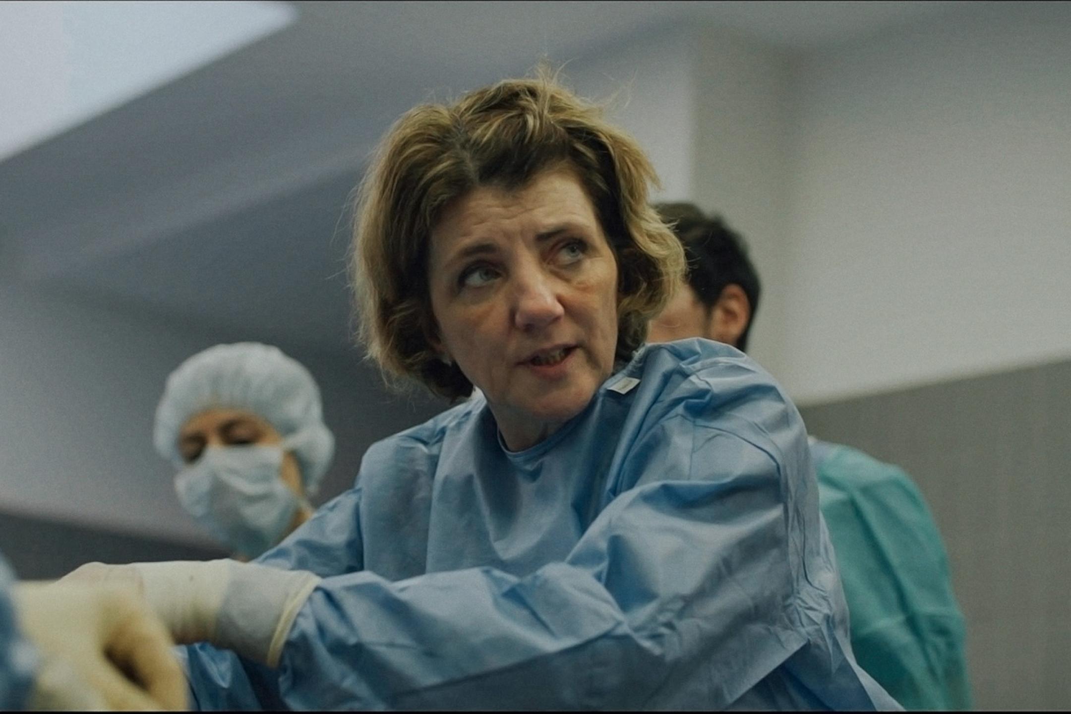 A female person in surgical dress stands leaning slightly forward, looking off to the side as she speaks.