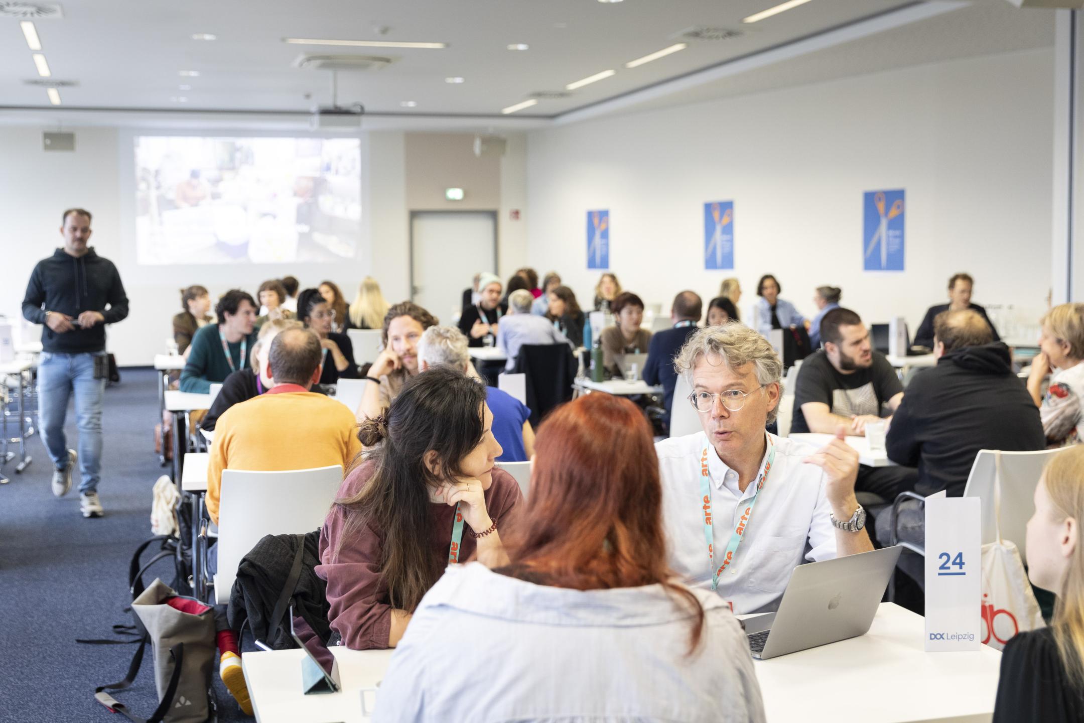 Viele Menschen sitzen in einem Raum an Vierer-Tischen, die in Reihen aufgestellt sind. Alle haben Laptops auf den Tischen und sind im Gespräch miteinander.