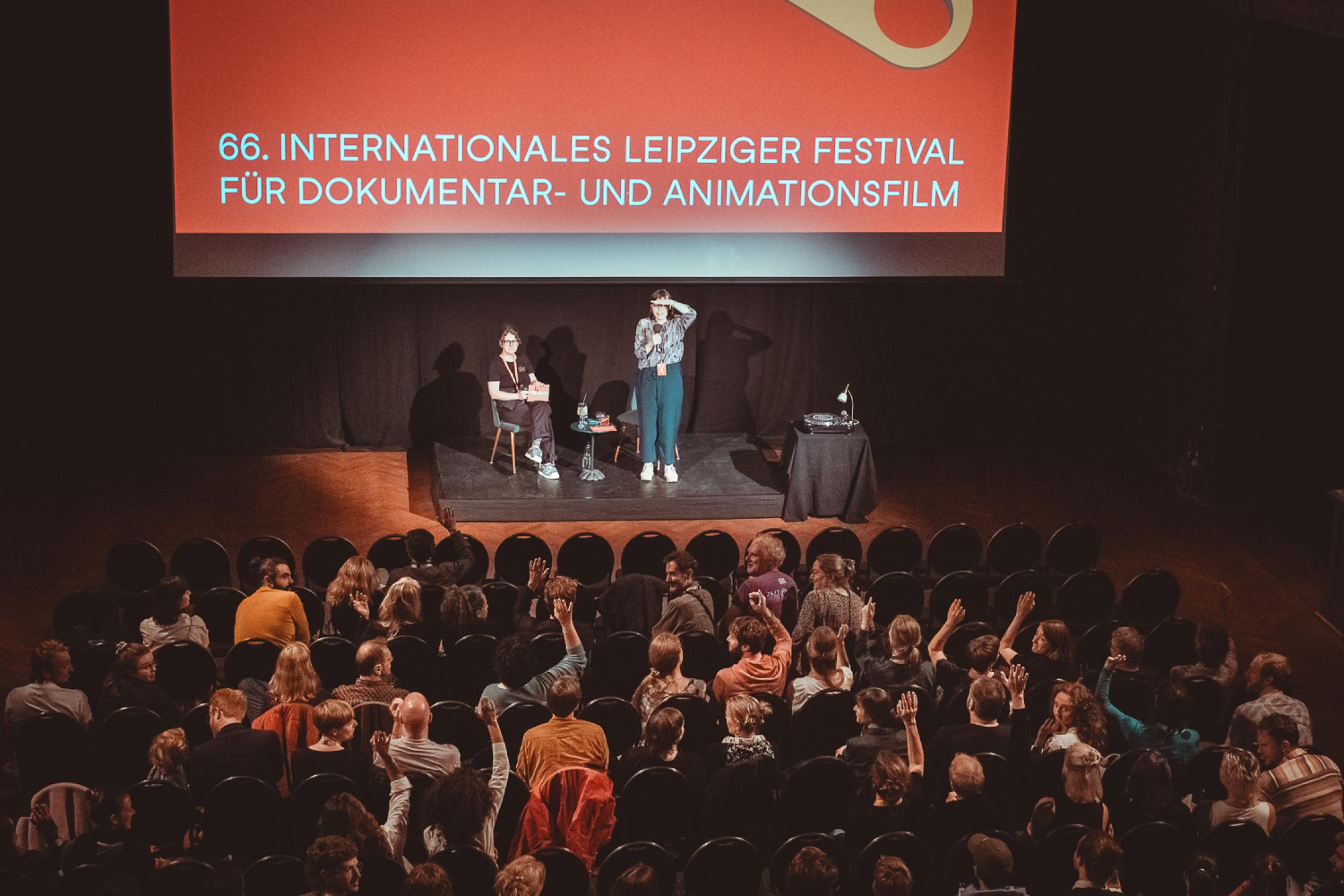 Auf einer Bühne vor einer Leinwand steht eine Frau mit Mikrofon und schaut in Richtung Publikum. Sie schirmt mit ihrer Hand das Licht von ihren Augen ab. Neben ihr sitzt eine zweite Person. Auf der Leinwand ist das Festivalmotiv von Dok Leipzig.