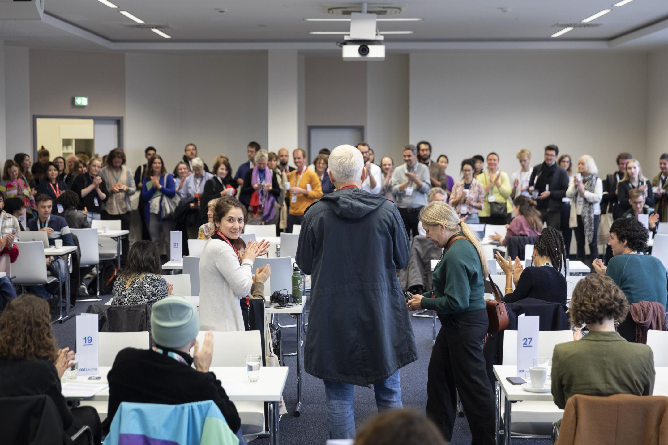 A conference room filled with a lot of people who stand and sit.