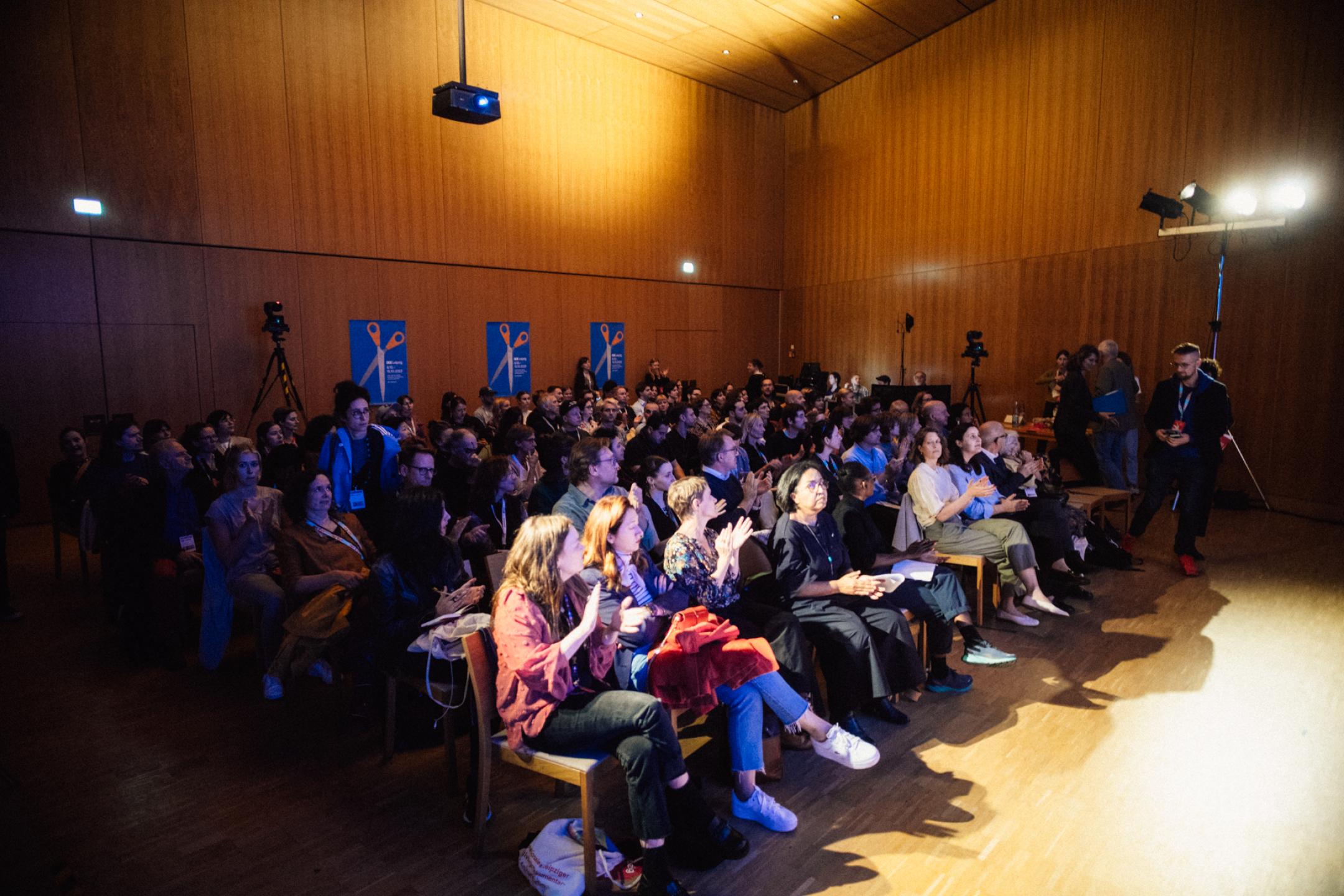 People sit in seat rows in a wood-paneled event room and are applauding. All seats are taken.