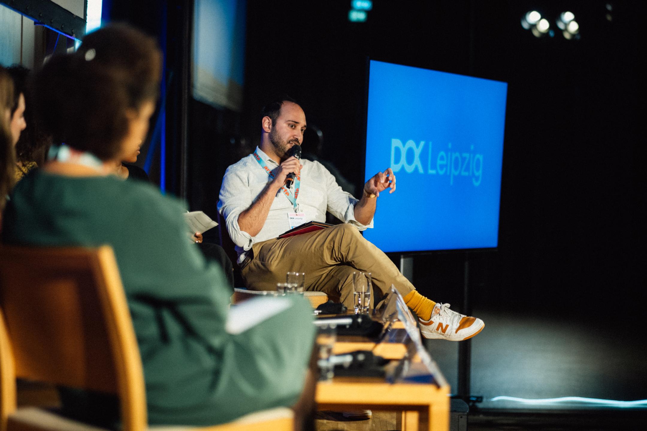 A man sits on a podium and speaks into a microphone. In the background a screen with the DOK Leipzig logo.