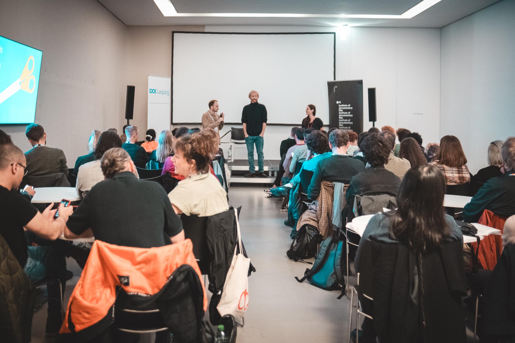 Three persons stand on a panel and test a microphone. The audience sit in seat rows and chat.