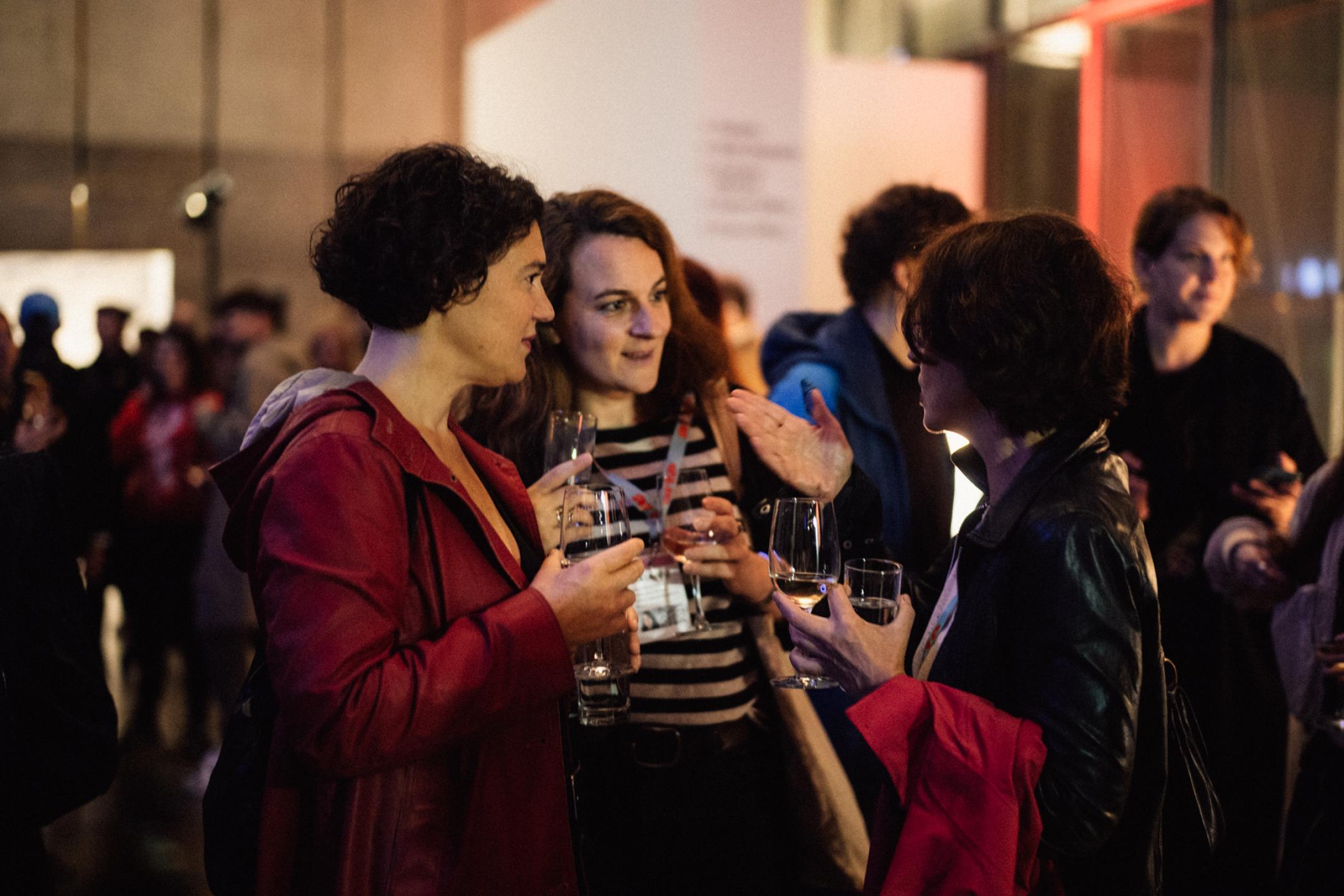 Three women stand together, holding glasses in their hands, and chat. Around them a lot of other persons.