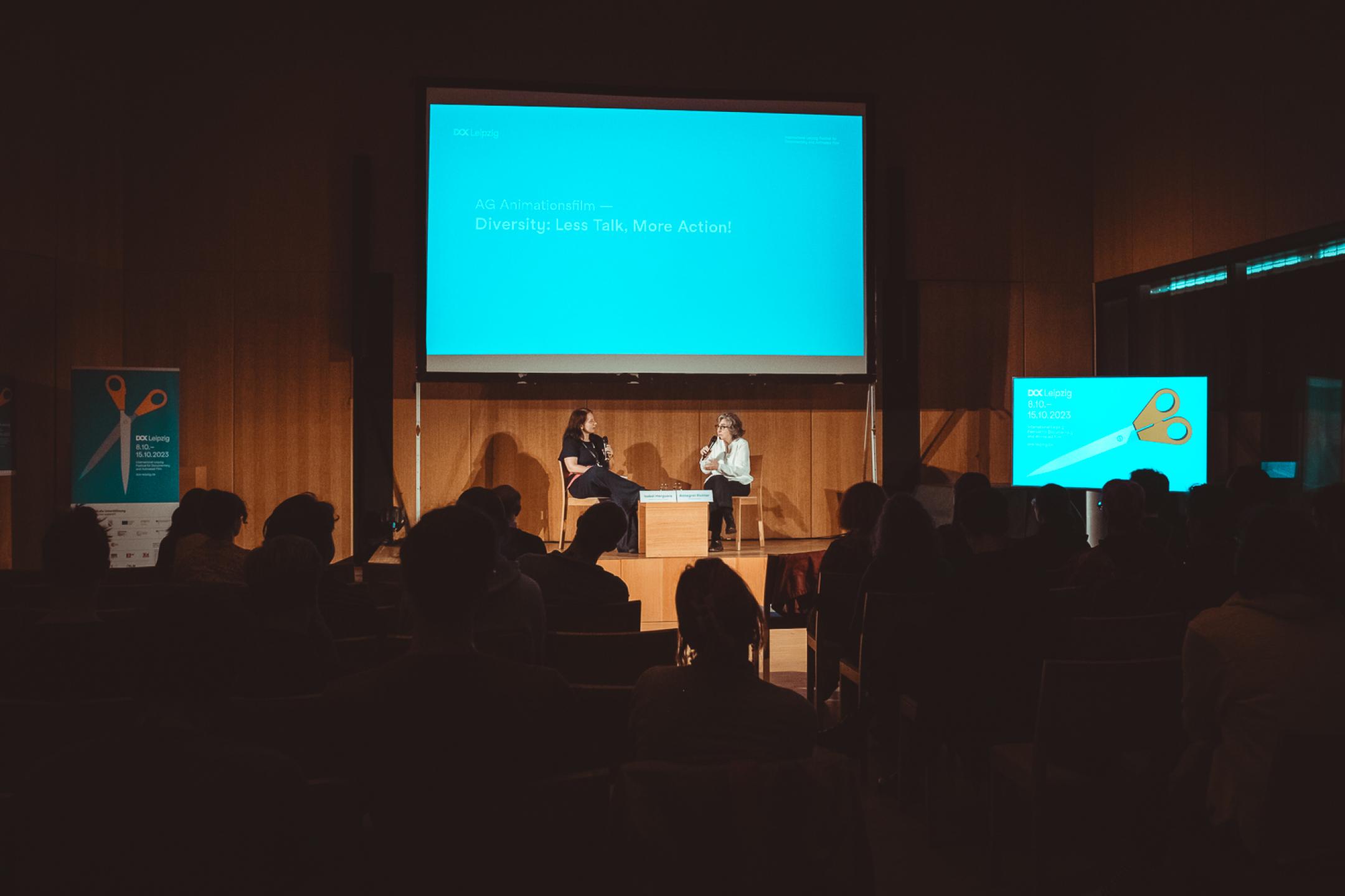 Two persons sit on a podium in front of an audience and talk to each other with microphones in their hands.