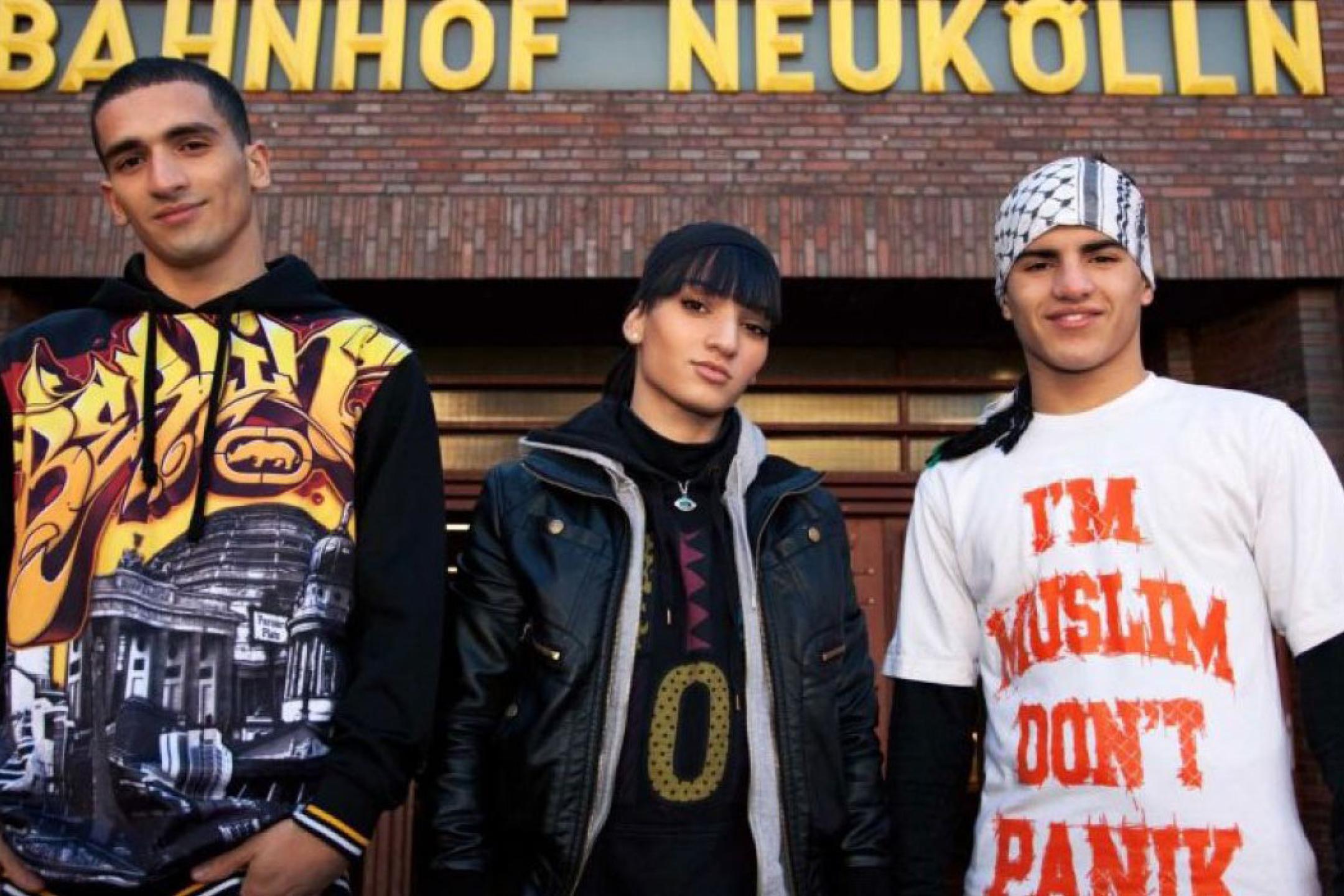 Drei junge Personen stehen vor dem Schild "Bahnhof Neukölln".