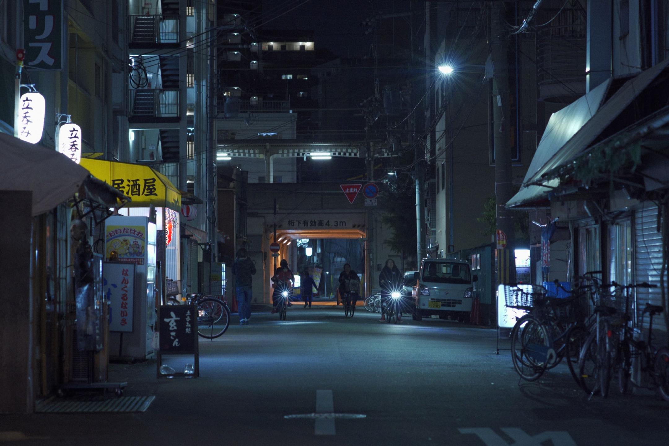 An empty alley at night with little shops and neon signs on the sides. Two persons on bicycles at the end of the alley.