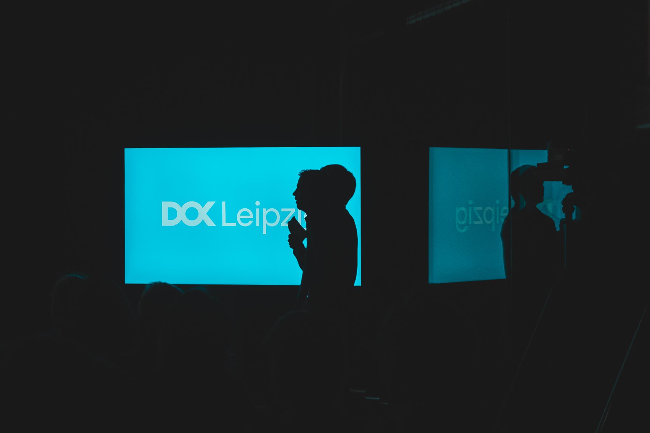 A dark room lit only by a blue screen with the DOK Leipzig logo. Two persons with microphones stand in front of the screen, only their shadows are visible.