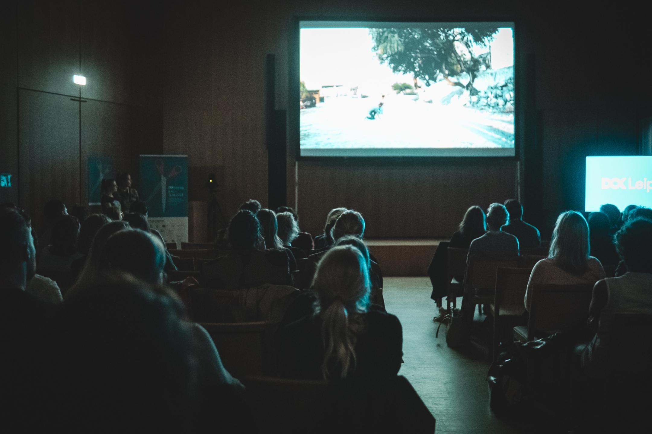 A dark room lit by a screen. A seated audience is looking at the screen.