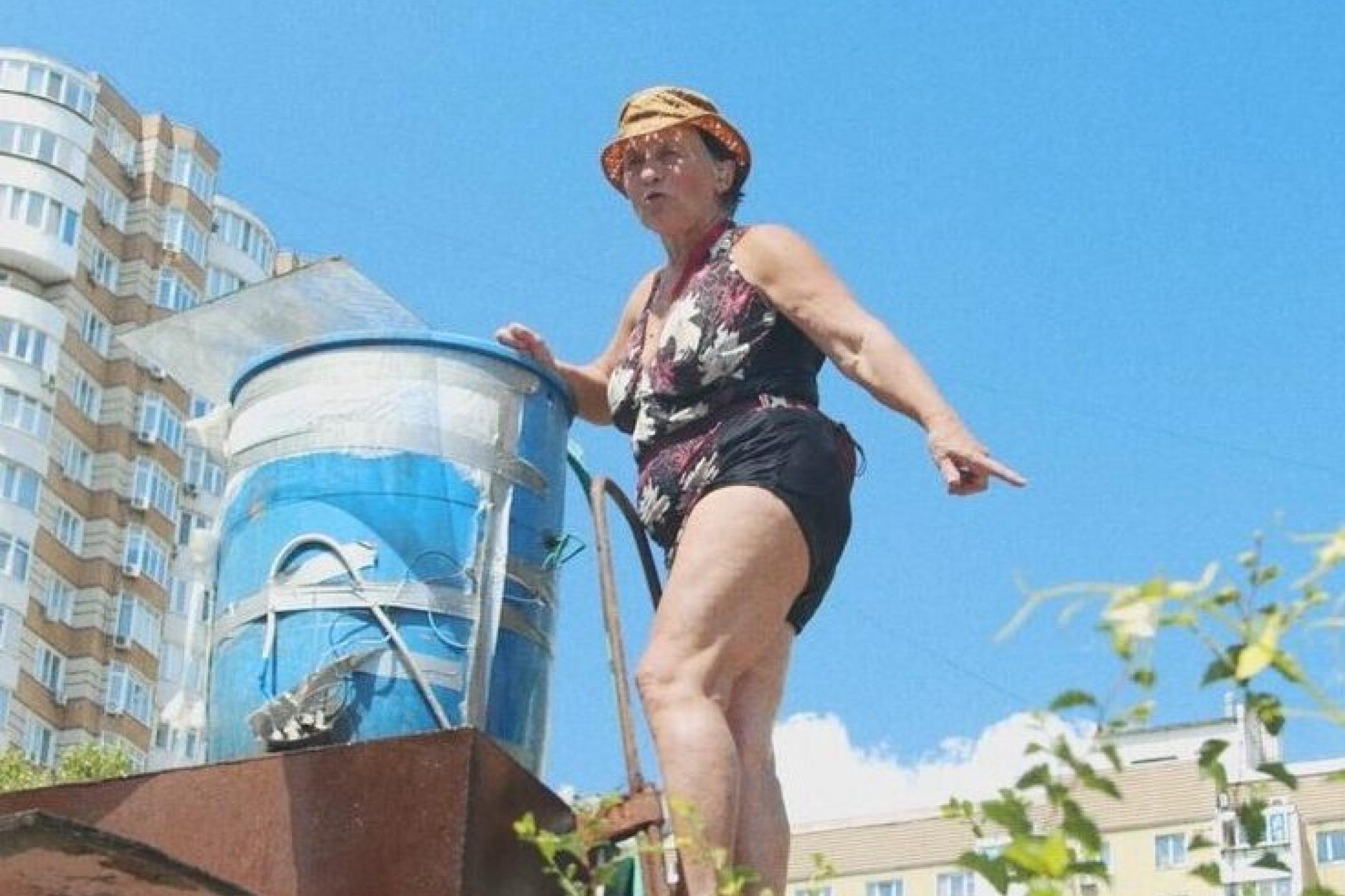 An old woman dressed in a bathing suit and a yellow hat is climbing a ladder towards a blue barrel that stands on a metal construction in a garden surrounded by urban houses