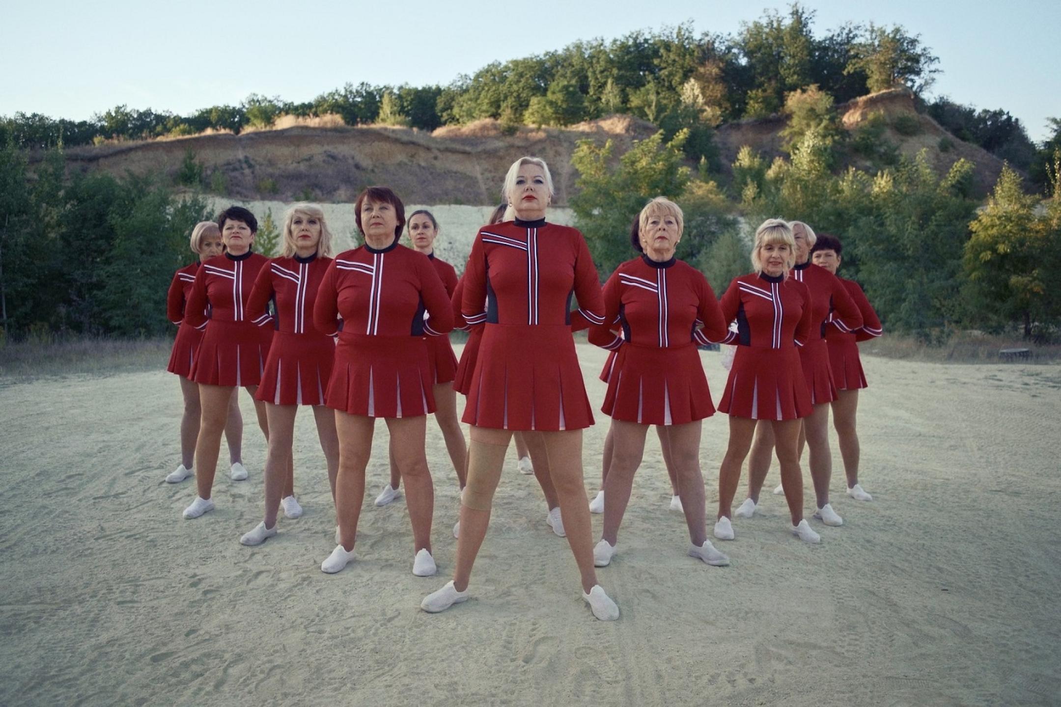 Eleven middle-aged women in read cheerleader uniforms are standing in position on a sandy field with their arms behind their backs and heads held high