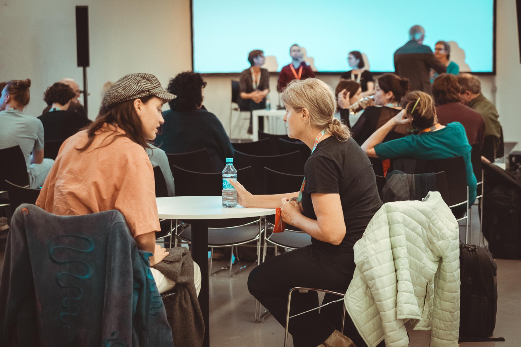 Two persons at a small round table talk while in the background panelists prepare for their talk