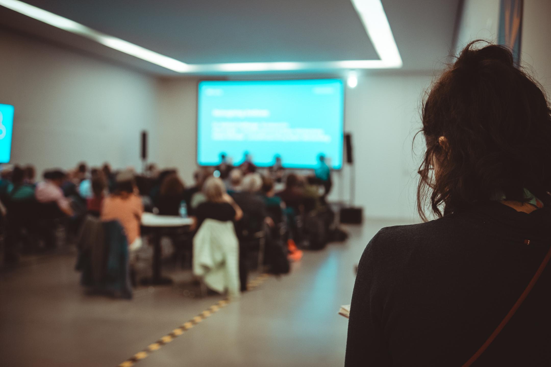 A person very close to the camera is looking into a room with an audience looking at a huge screen in industry blue