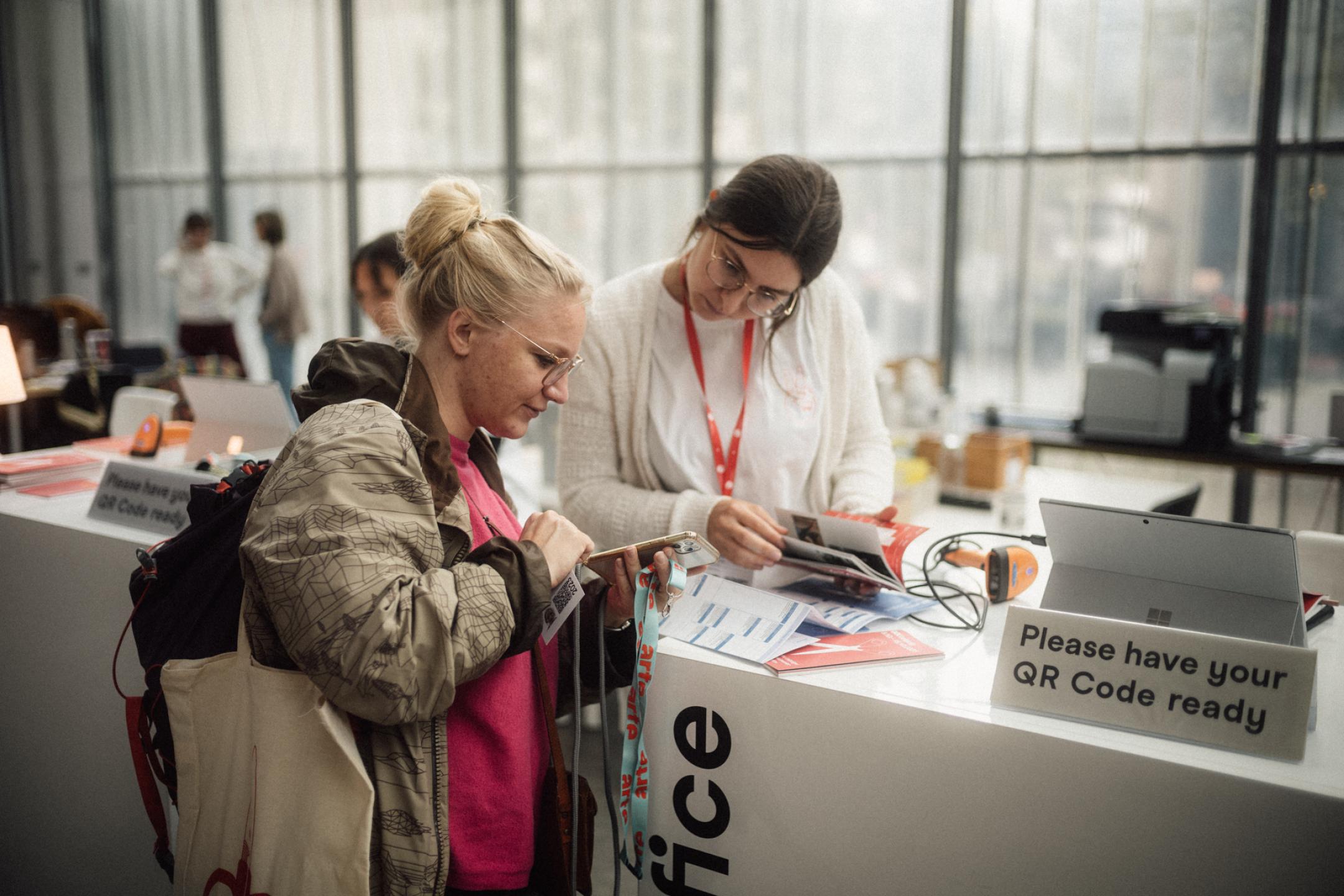 zwei Personen stehen an einem Tisch, beide schauen nach unten auf Flyer, Programmhefte und ein Smartphone, das eine Person in der Hand hält