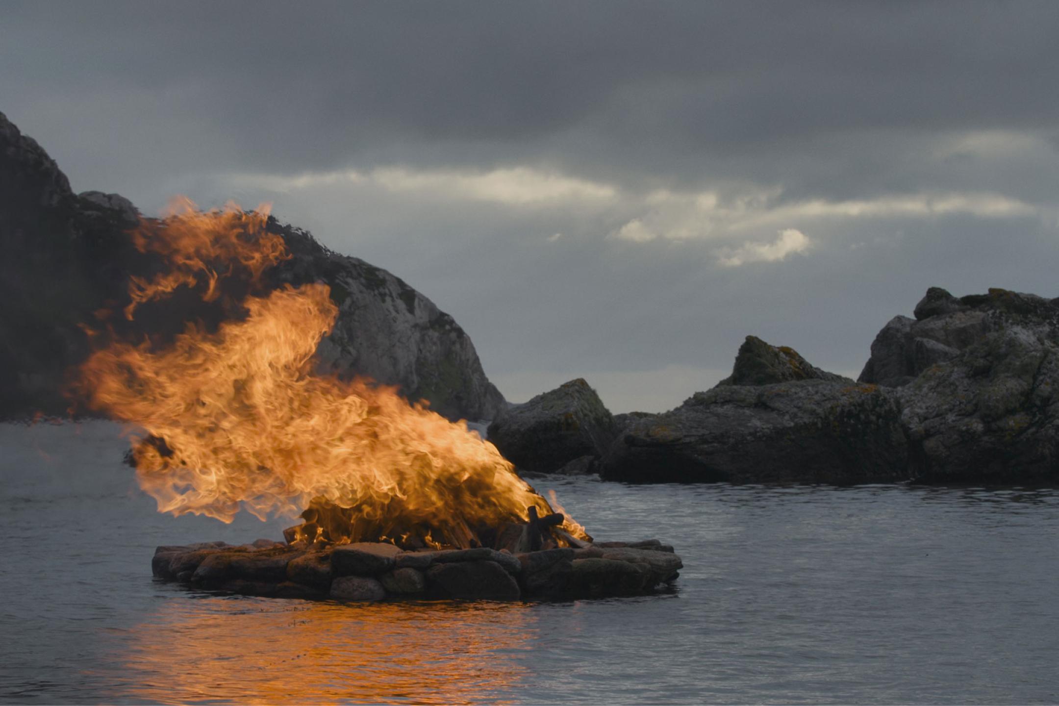 A film still from "Tracing Lights", showing a stack of wood burning brightly on a lake, while dark clouds and distant mountains loom over the scenery.