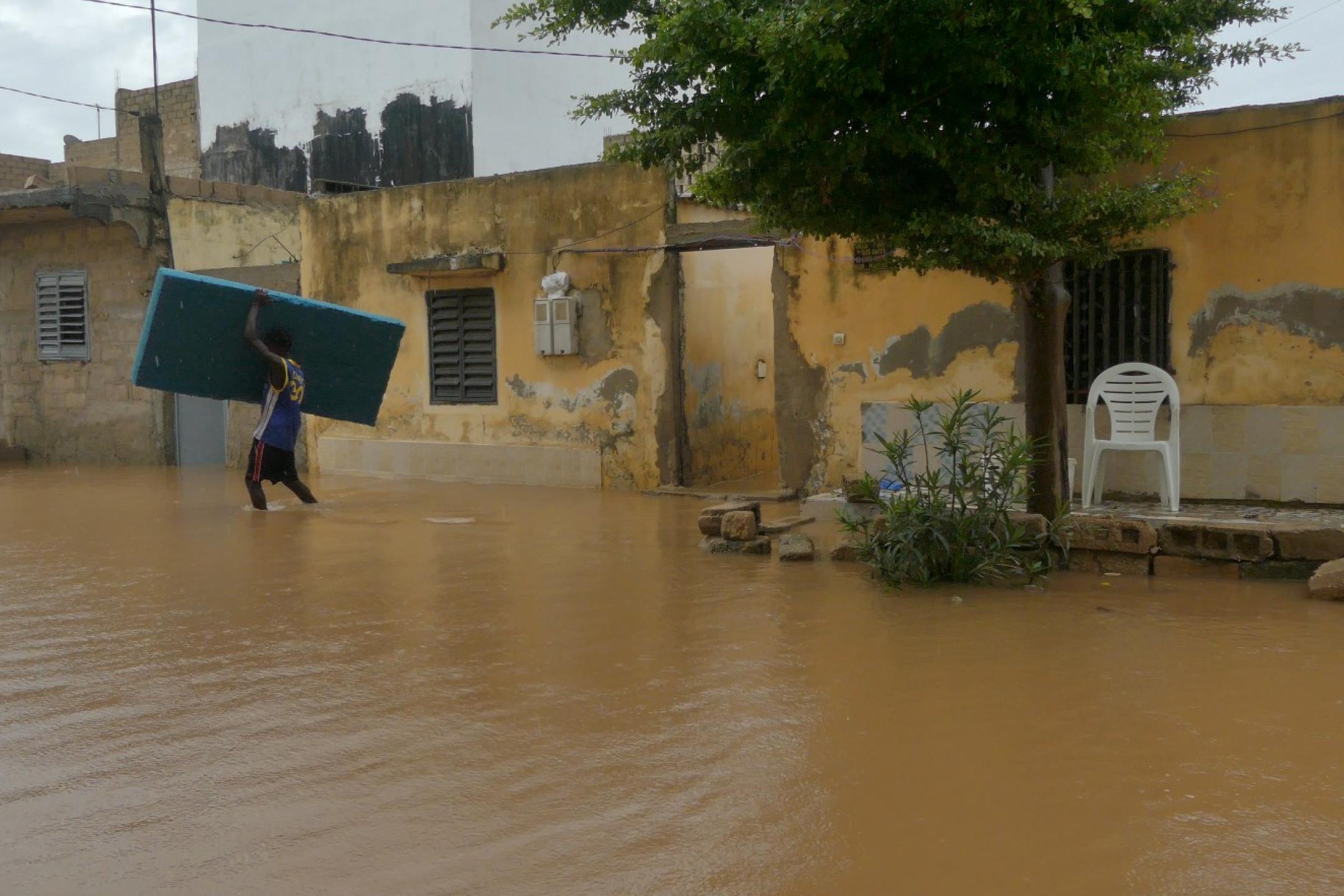 A man is carrying a mattress across a flooded road next to little yellow houses