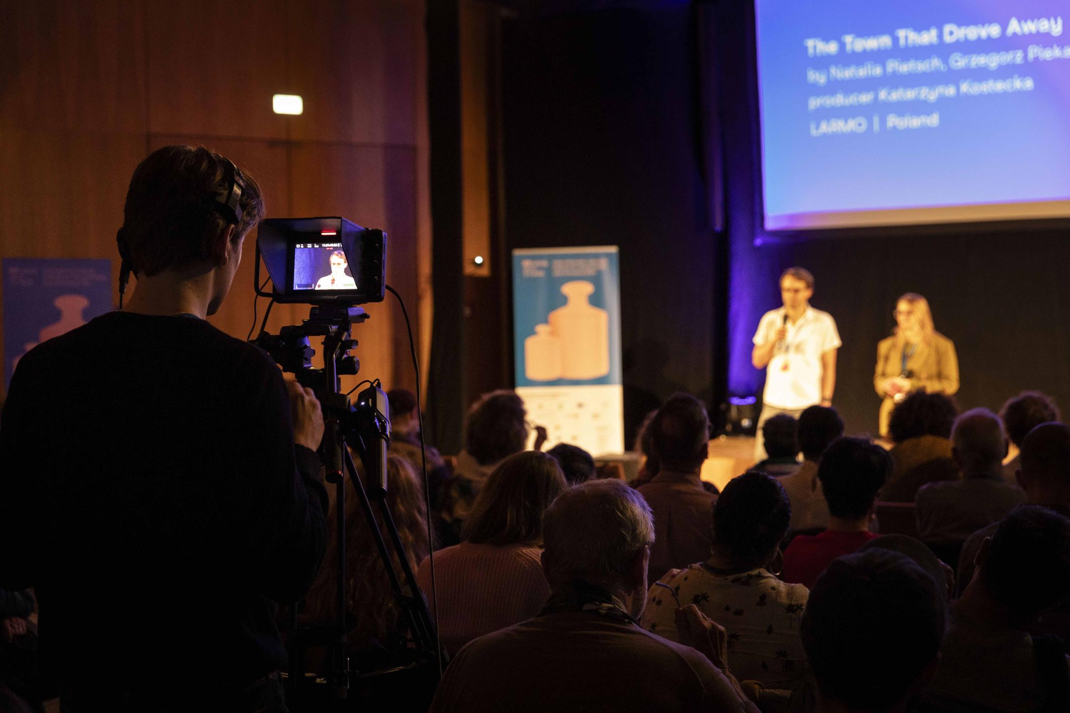 Two people on stage pitch their project to an audience, a cameraman is filming them