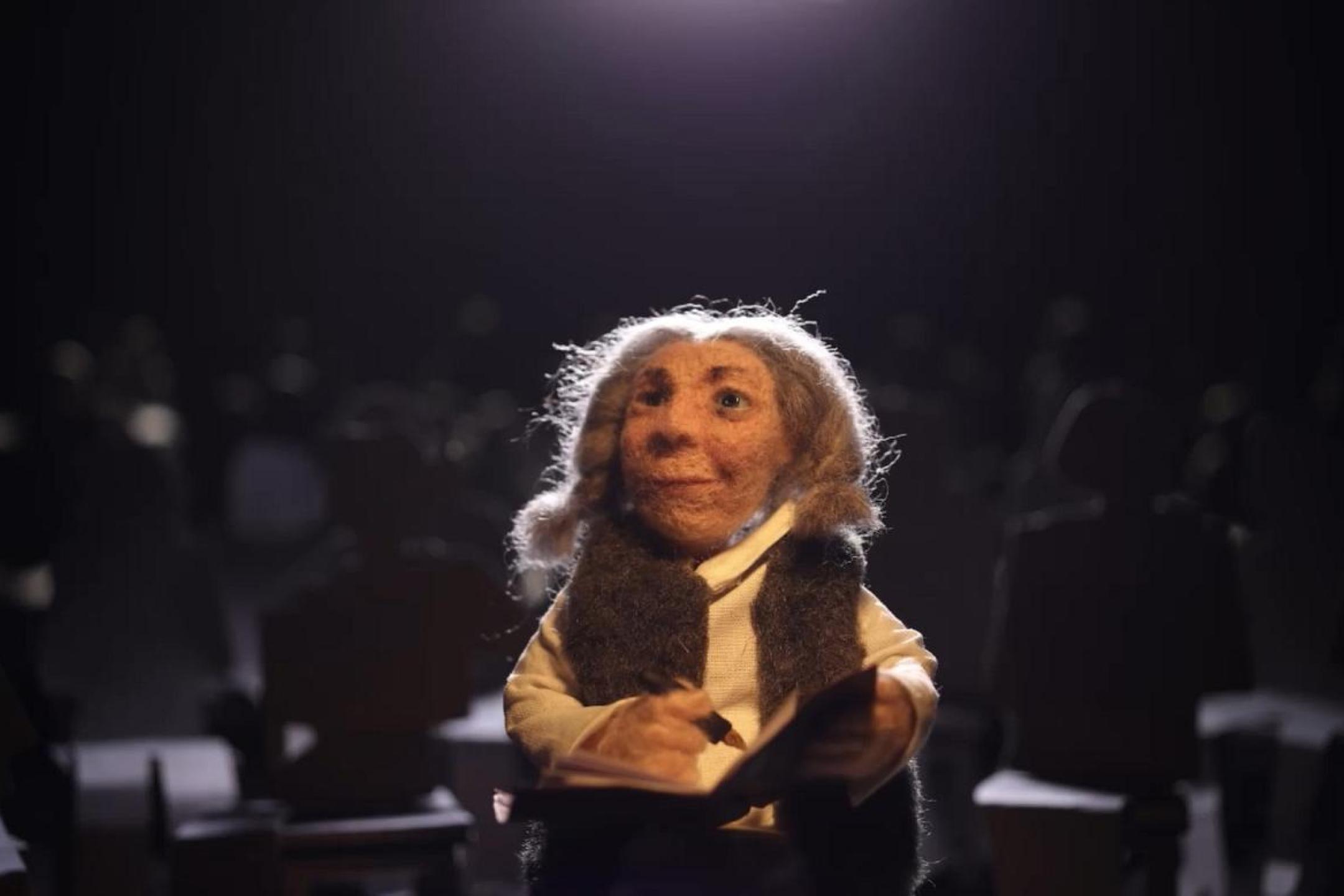 An animated puppet of a grey-haired woman is standing in a theater hall writing in a book