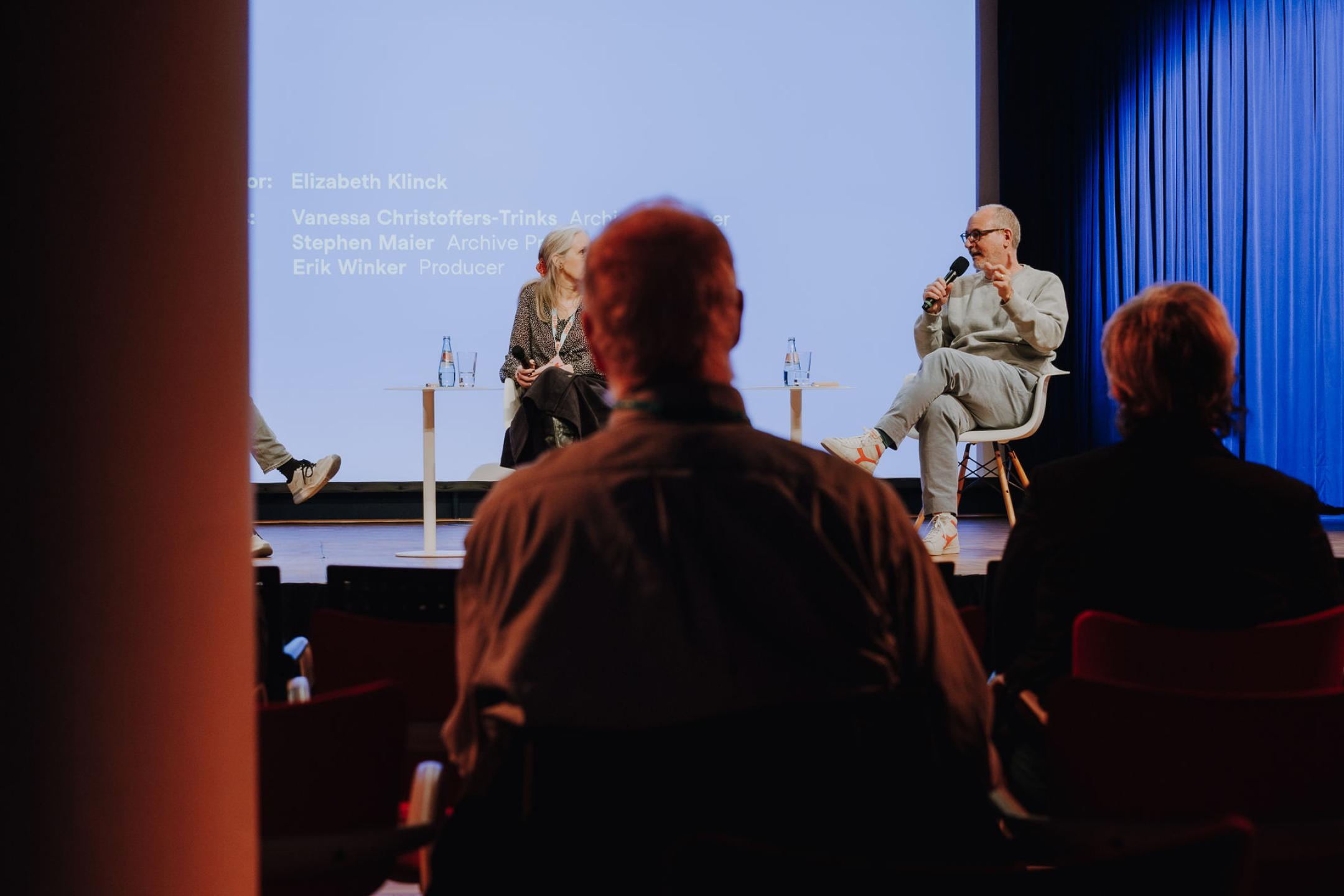 photograph of a man sitting in an audience watching people talk on a stage