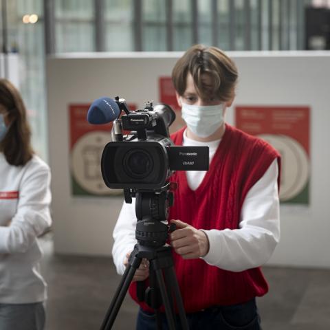 Ein Jugendlicher positioniert eine semi-professionelle Kamera mit Aufsteckmikrofon und Stativ im Festivalzentrum von DOK Leipzig. Im Hintergrund mehrere DOK-Plakate.