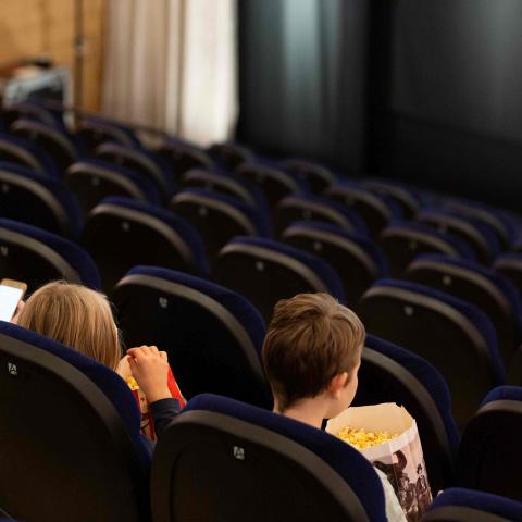 Im Kino: Drei Kinder sitzen mit Popcorn-Bechern auf ihren Sitzplätzen in einer der vorderen Reihen. Sie warten darauf, dass der Film beginnt.