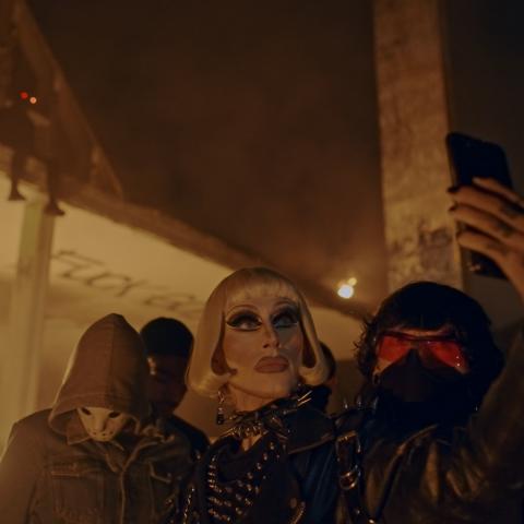 In the center you can see a heavily made-up person with striking facial features. She wears a white-blond wig with a bob cut and black patent clothing. She is taking a selfie with a serious expression. To her left and right are people wearing face masks and hoods. Everything looks a little sinister, but also rebellious and powerful.