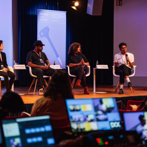 Vier Personen (zwei Frauen, zwei Männer) sitzen an einer Tafel. Der Mann auf der rechten Seite spricht in ein Handmikrofon, die anderen hören zu. Vorne leuchten die Monitore eines Live-Schnittplatzes in der Dunkelheit auf.