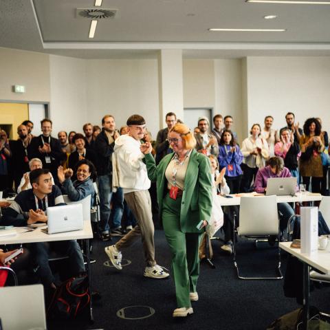 In a large meeting room: A woman and a man are holding hands, beaming with joy. Around them is a group of many people applauding.