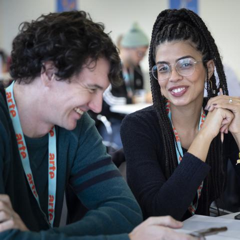 A woman of colour and a white man, both laughing, are sitting at a table at the Co-Pro Market