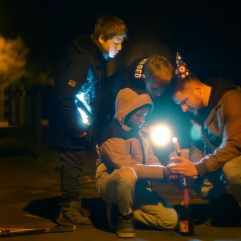 Auf einer dunklen Straße zünden ein Kind und ein Mann mit Partyhut eine Silvesterrakete. Zwei weitere Kinder schauen neugierig zu