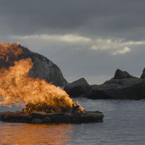 A film still from "Tracing Lights", showing a stack of wood burning brightly on a lake, while dark clouds and distant mountains loom over the scenery.