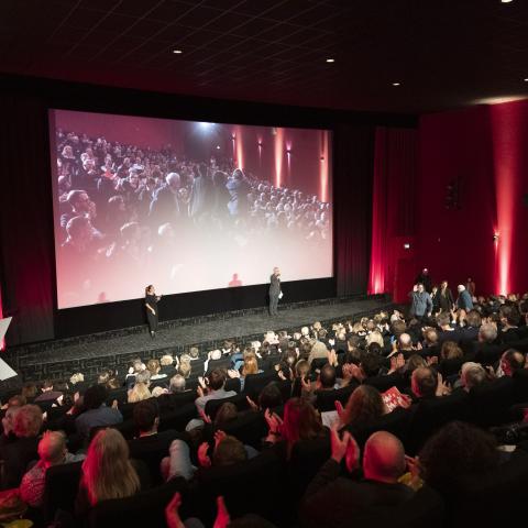 Das Bild zeigt die Eröffnung des Festivals im Kino: das Publikum im Anschnitt, die Leinwand, die Bühne davor mit Moderatorin und Festivalleiter