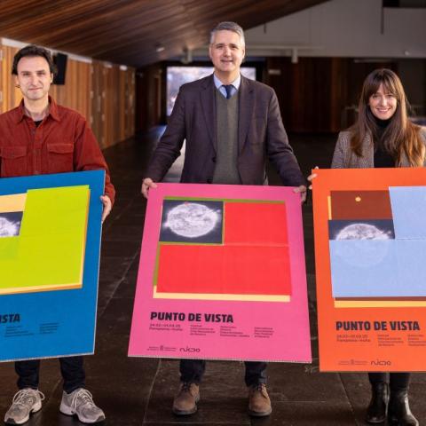 Two men and a woman stand smiling in front of a brown wooden wall. They are holding colourful festival posters.