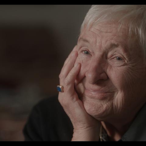 An elderly woman looks into the camera, her head resting in her hand, smiling thoughtfully
