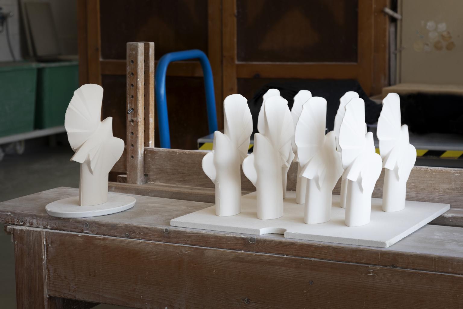 On a wooden table covered with white dust, a dozen white porcelain doves are lined up. To their left is another one, greyish and a little rougher.