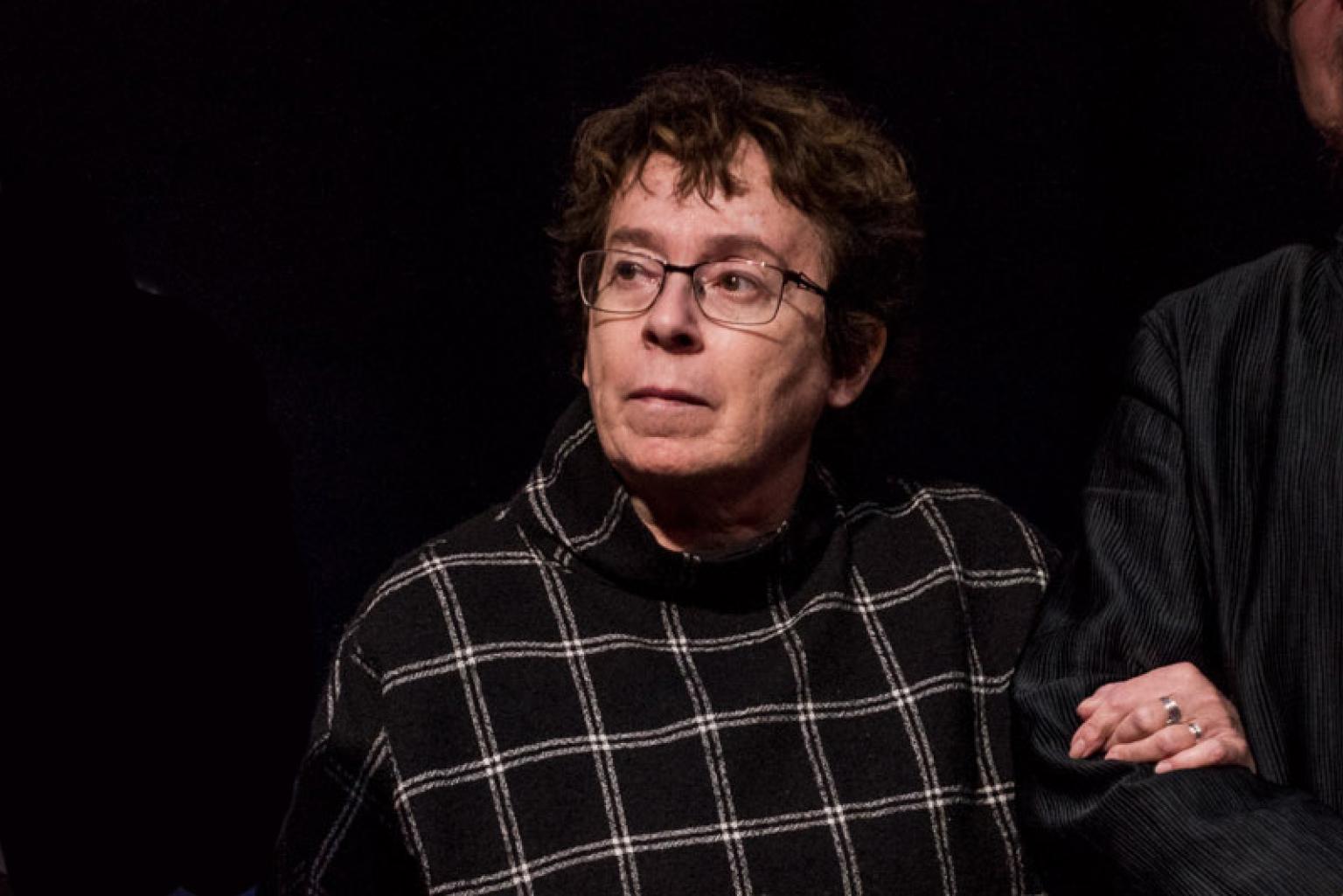 Portrait of Heddy Honigmann. She stands on a stage, looking attentively towards the audience while listening.