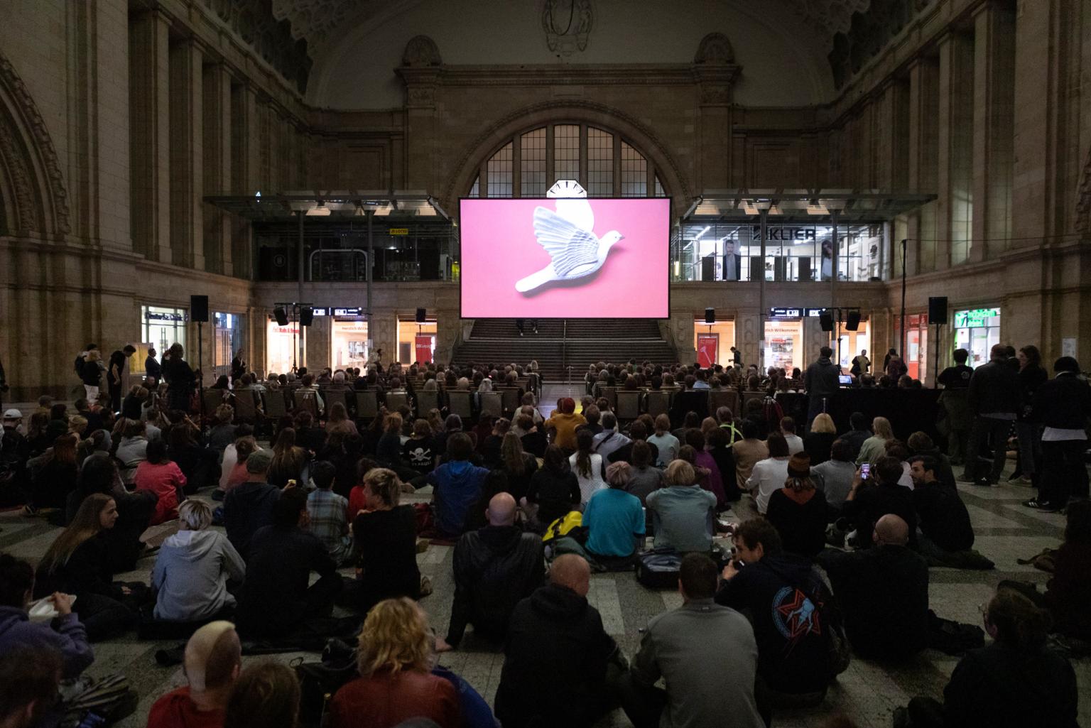 In einer großen Halle ist eine Kinoleinwand mit Sitzreihen aufgebaut. Die Sitzreihen sind voll besetzt, rundherum und dahinter sitzen zahlreiche Zuschauer*innen auf dem Boden. Auf der Leinwand fliegt eine weiße Taube auf pinkem Hintergrund.