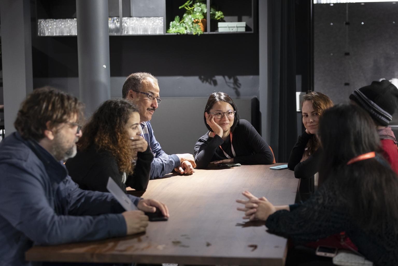 Several persons (women and men) are sitting around a big table. One person is talking, the others are listen and smile.