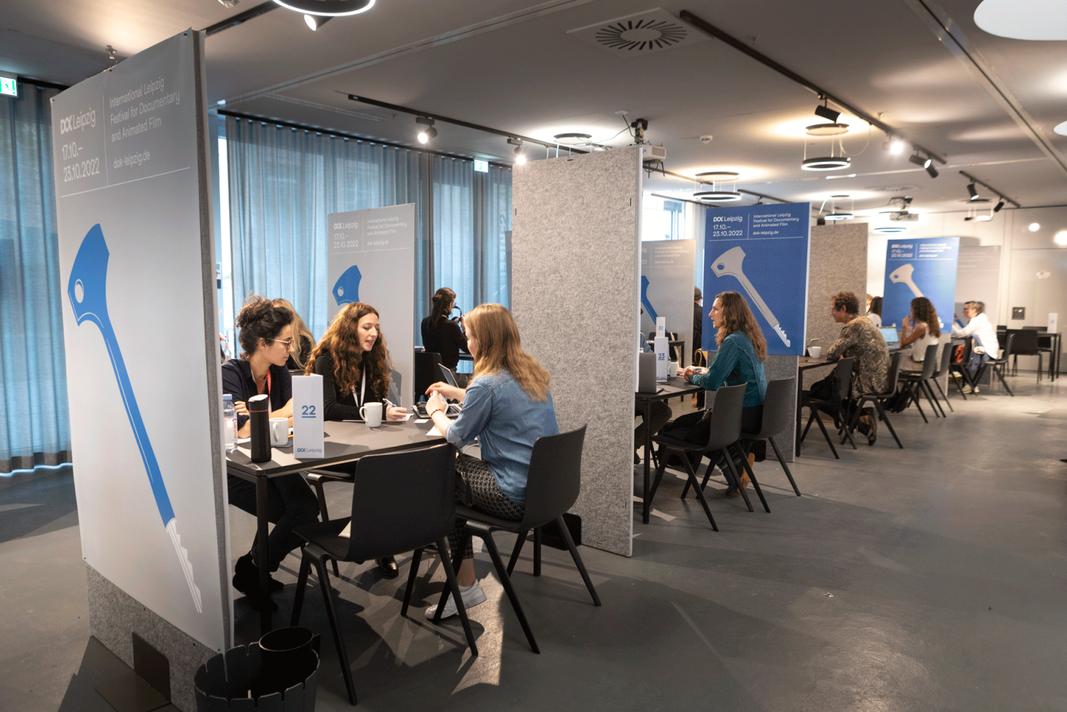 A set of conference tables separated by partitions. Three to four persons are sitting at each table talking to each other.