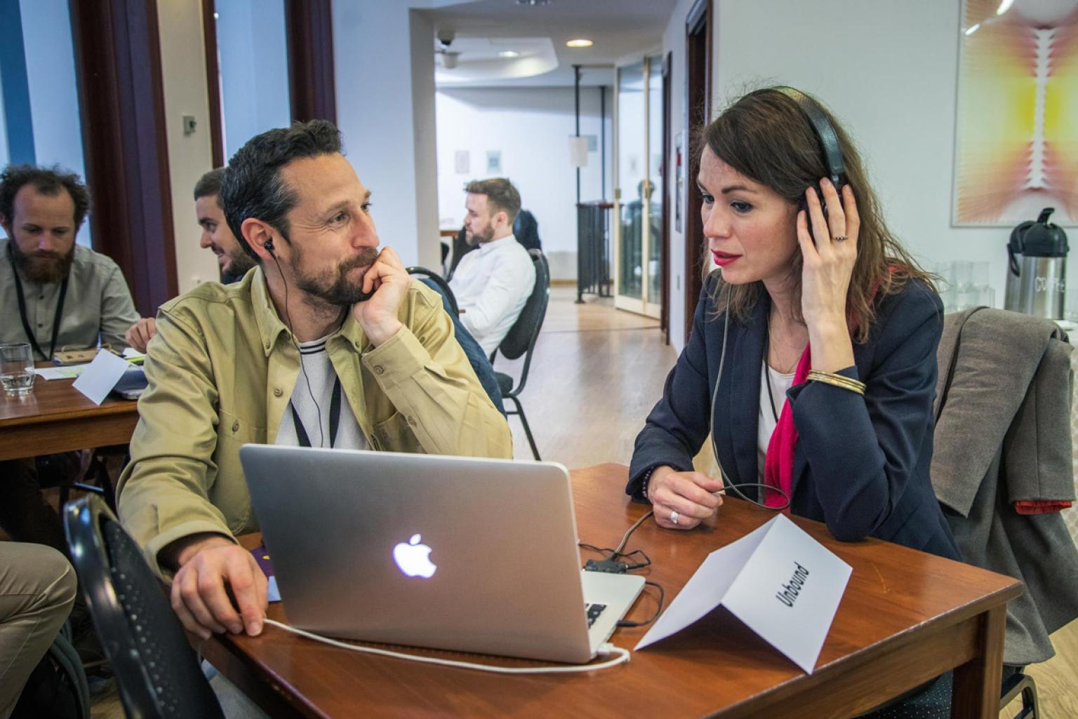 Two persons sit at a small table, the woman (at the right) wears earphones and looks at a laptop. The man (at the left) watches her.