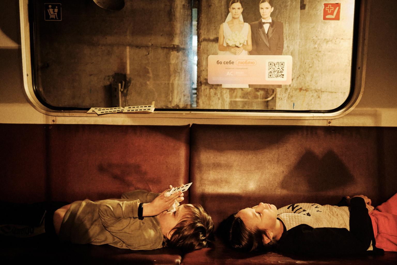 Two children lying relaxed on a bench of a standing subway car. A view out of the carriage window reveals the wall of a subway tunnel.