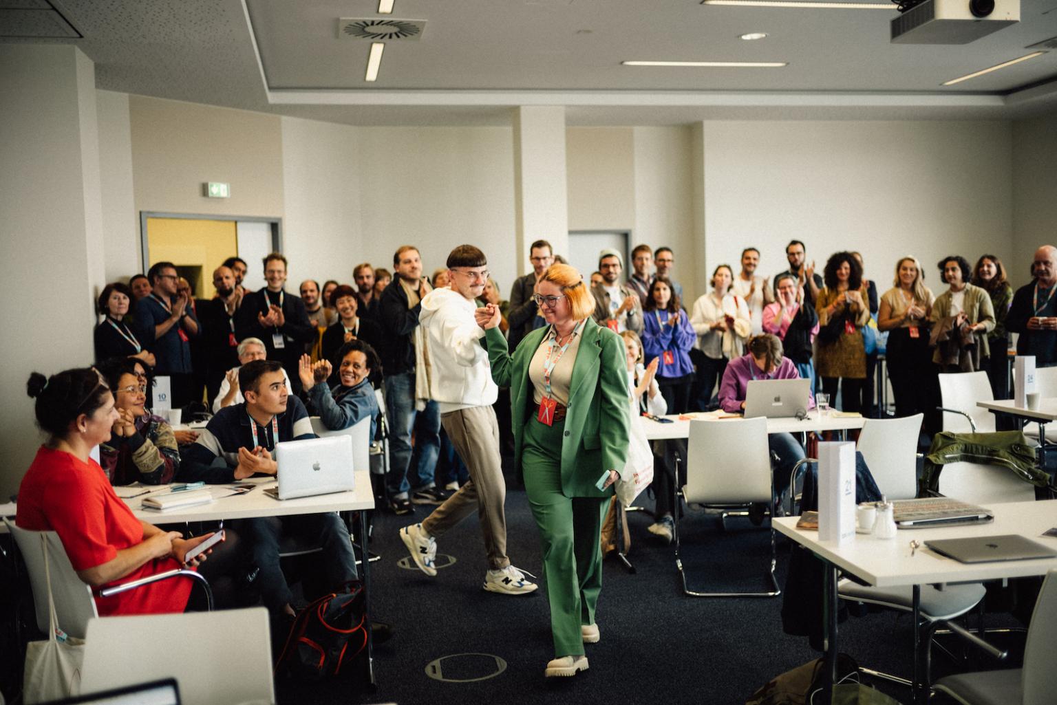 In a large meeting room: A woman and a man are holding hands, beaming with joy. Around them is a group of many people applauding.