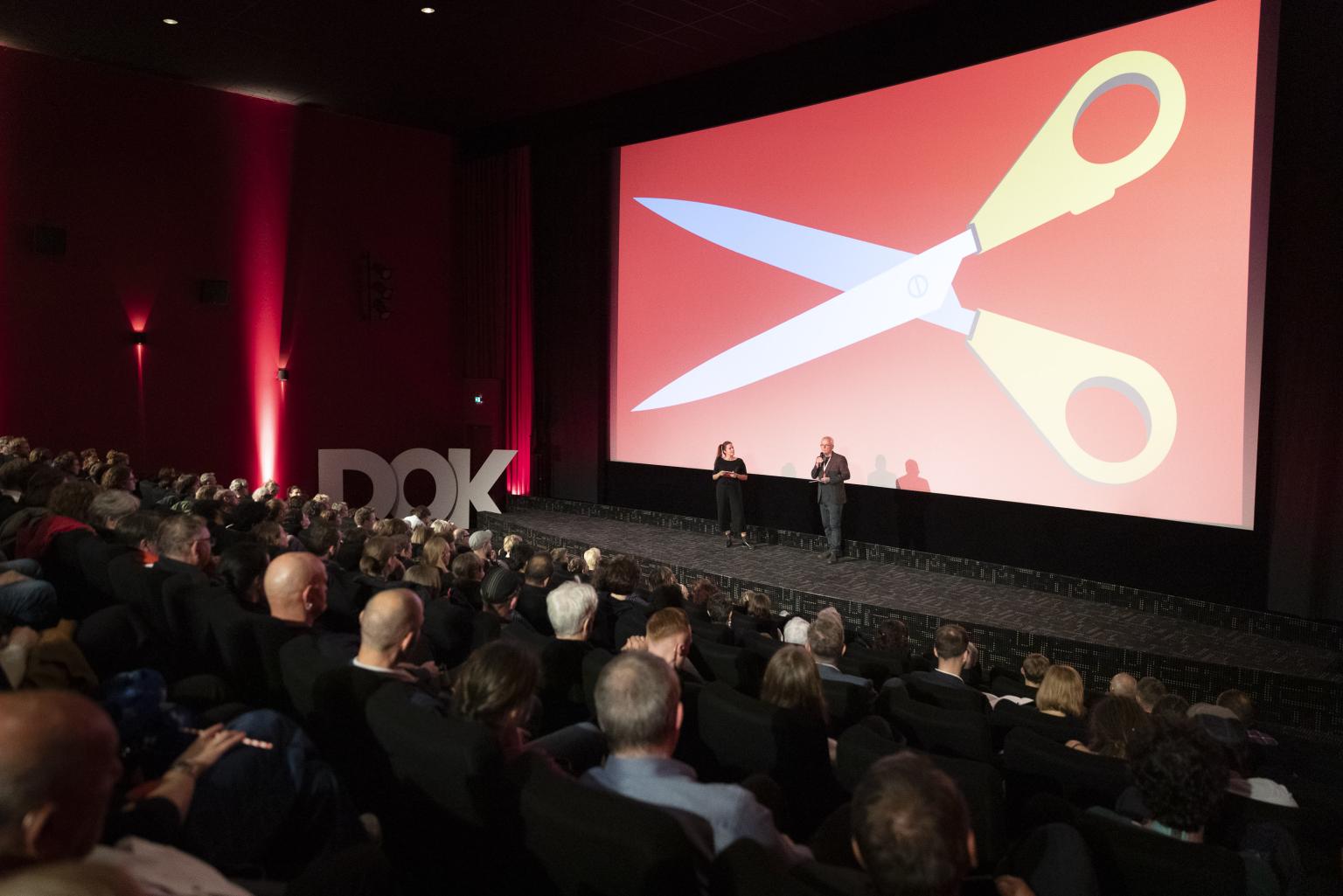 Festivaldirektor Christoph Terhechte und Moderatorin Julia Weigl auf der Bühne bei der Festivaleröffnung 2023. Auf der Leinwand hinter ihnen ist eine riesige Schere abgebildet.